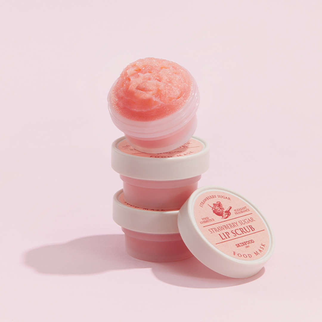 A stack of SKINFOOD STRAWBERRY SUGAR FOOD MASK LIP SCRUB containers on a pastel pink background. The top container is open, revealing a textured, sugary pink scrub. Another container lies on its side, showing the “Strawberry Sugar Lip Scrub” label clearly.