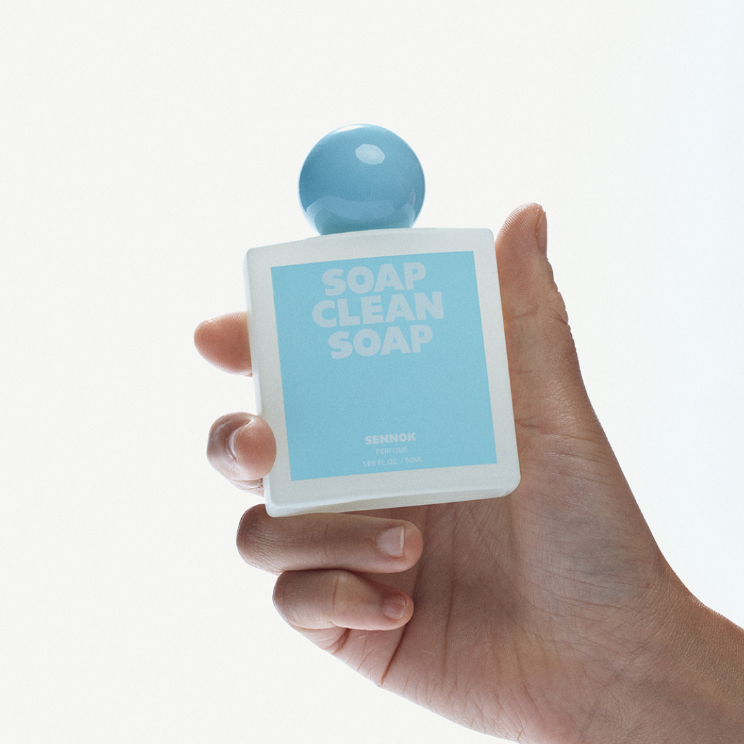 A close-up of a hand holding a square bottle of SENNOK “Soap Clean Soap” perfume, featuring a light blue label and a round blue cap against a bright, minimal background.