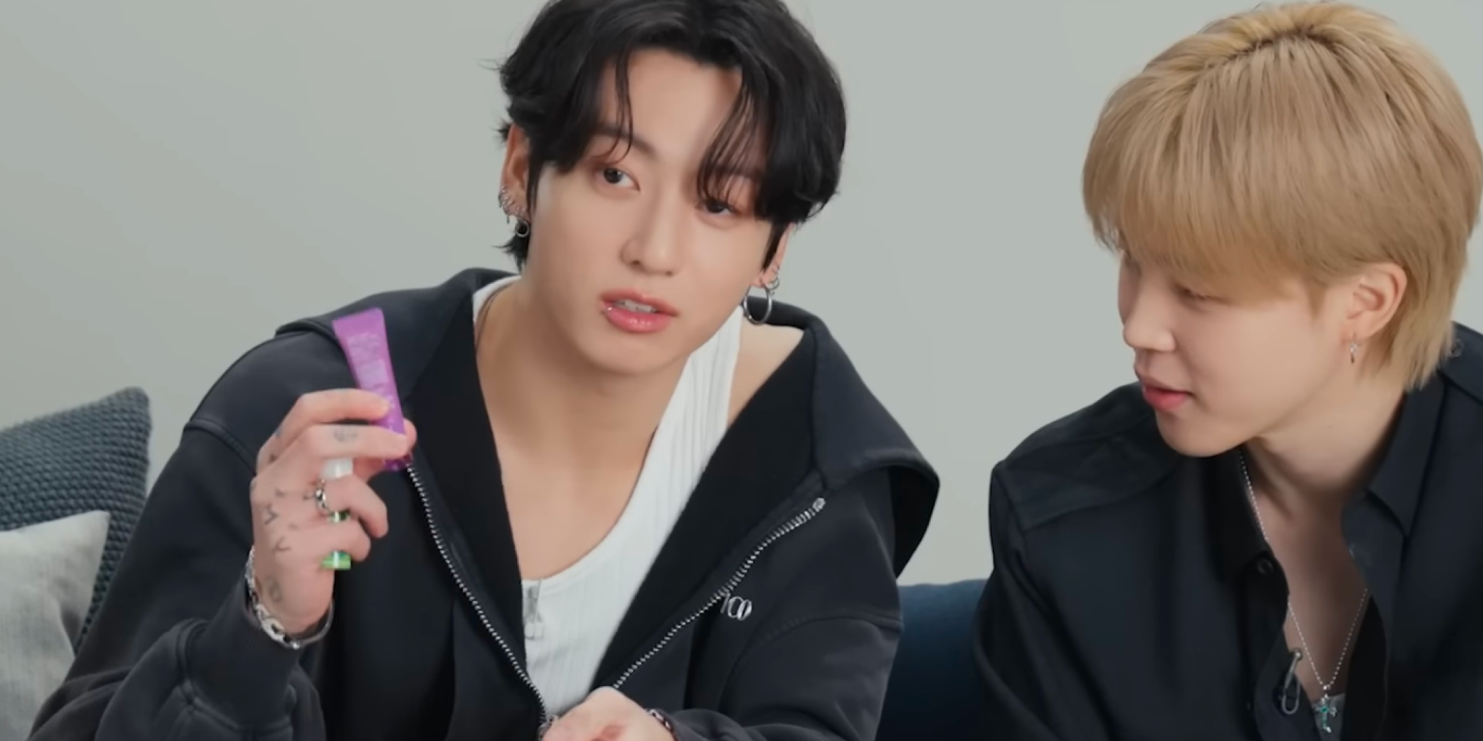 A BTS member, Jungkook, appears on a Vogue Japan YouTube segment holding up a purple tube of LANEIGE Lip Glowy Balm in the Gummy Bear scent while speaking. Another member sits beside him, watching attentively as they chat together on a sofa in a relaxed setting.