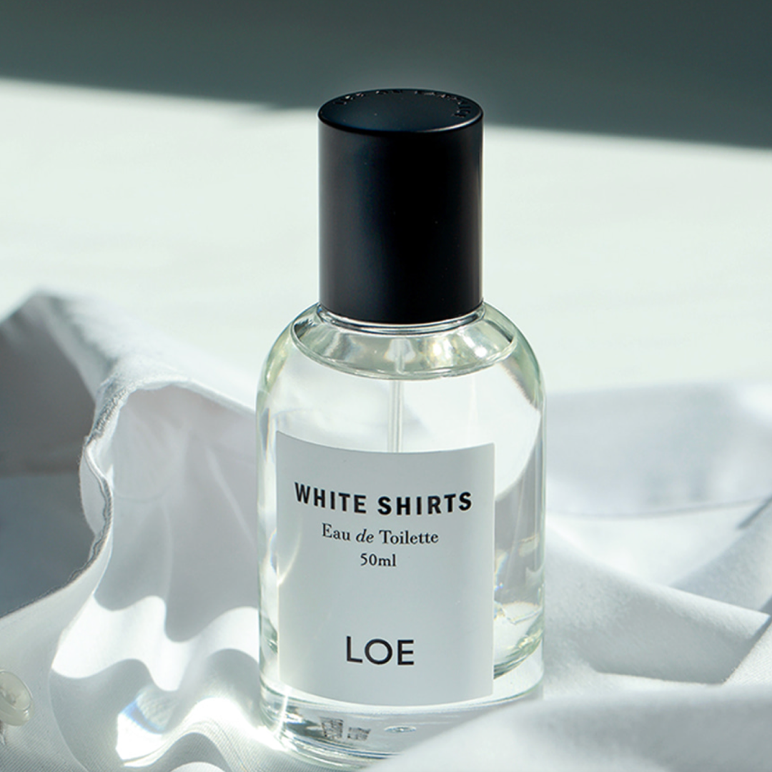 A clear glass bottle of LOE’s “White Shirts” Eau de Toilette sits on softly folded white fabric, illuminated by natural sunlight for a clean and fresh aesthetic.