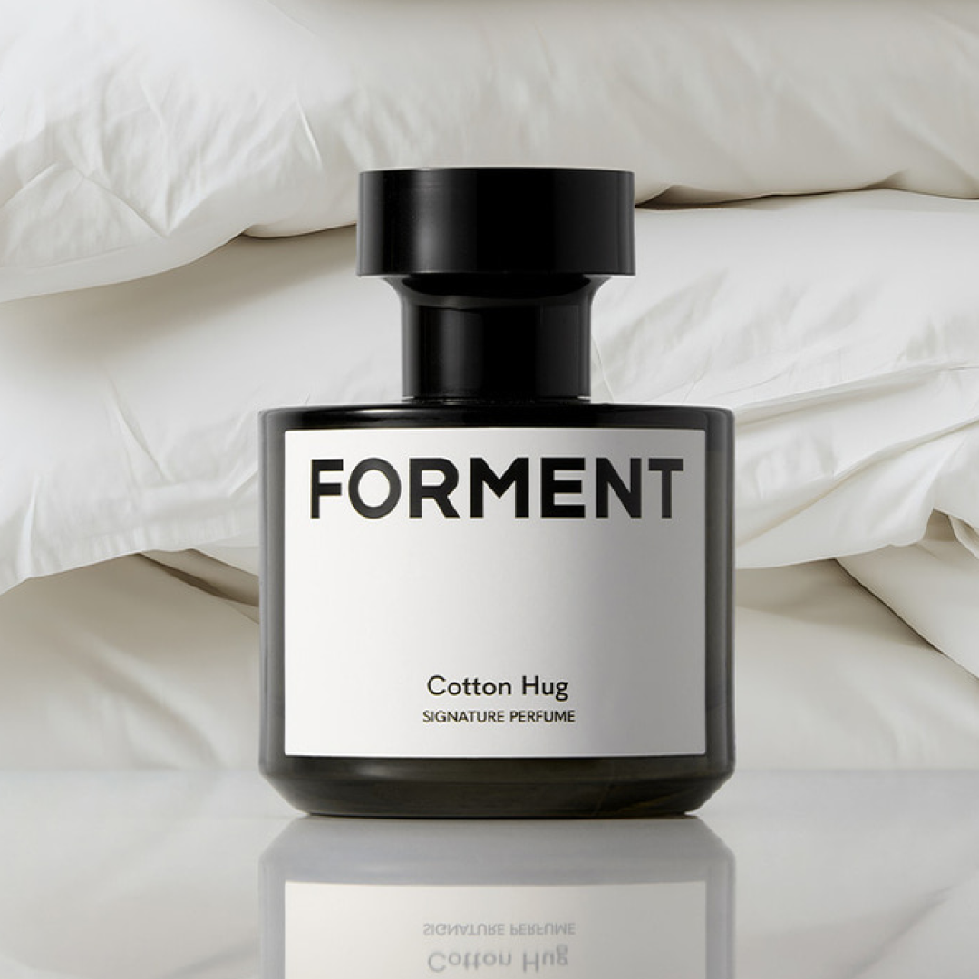 A black perfume bottle labeled “Forment Cotton Hug” is placed in front of neatly stacked white bedding, creating a clean and cozy visual mood.