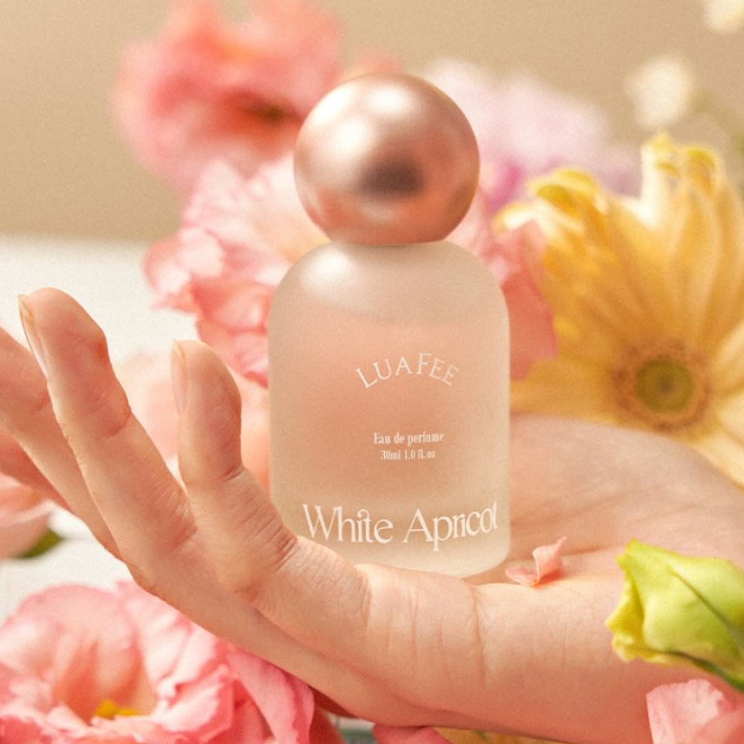 A hand gently cradles a frosted bottle of LUAFEE White Apricot Eau de Parfum, surrounded by soft pink and yellow flowers in a warm, dreamy setting.