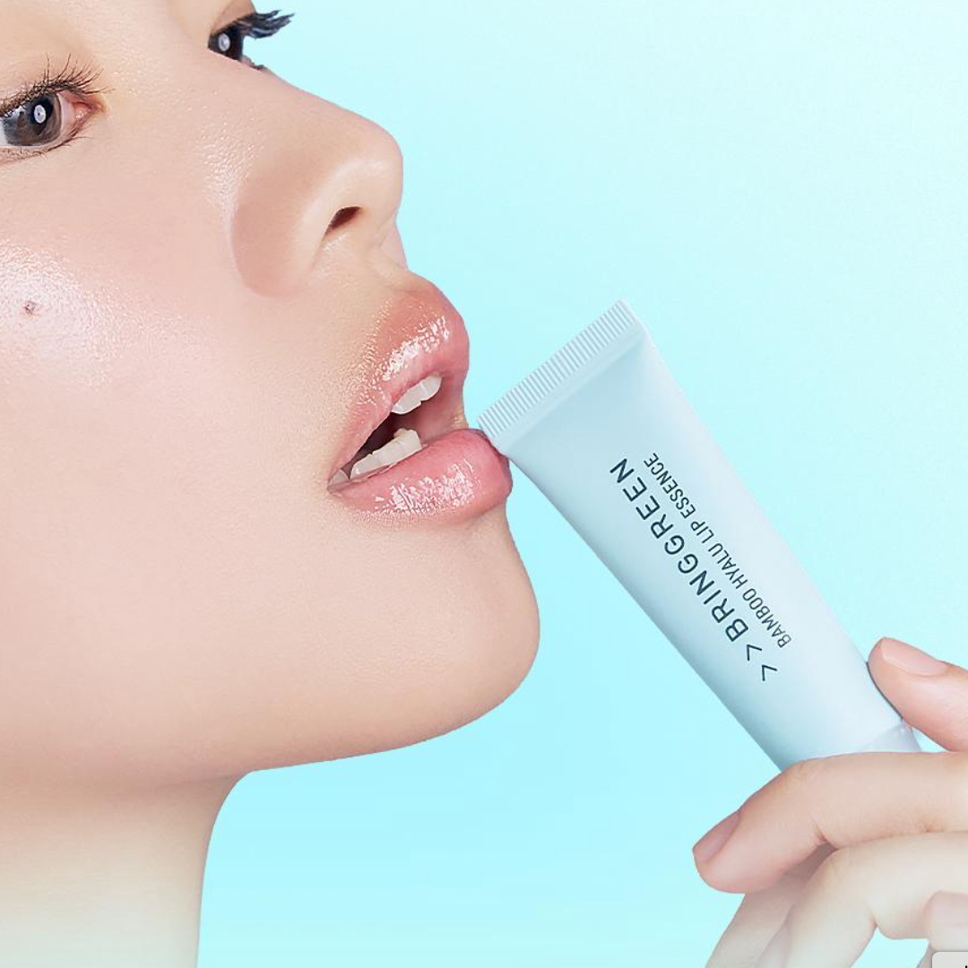 A close-up of a model applying a glossy lip essence from a light blue BRINGGREEN Bamboo Hyal Lip Essence tube. The model’s lips appear smooth, hydrated, and shiny against a clean sky-blue background.